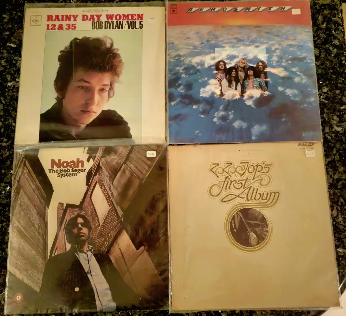 Four vintage vinyl record album covers are arranged in a grid on a dark countertop. Top left: Bob Dylan's 'Rainy Day Women…