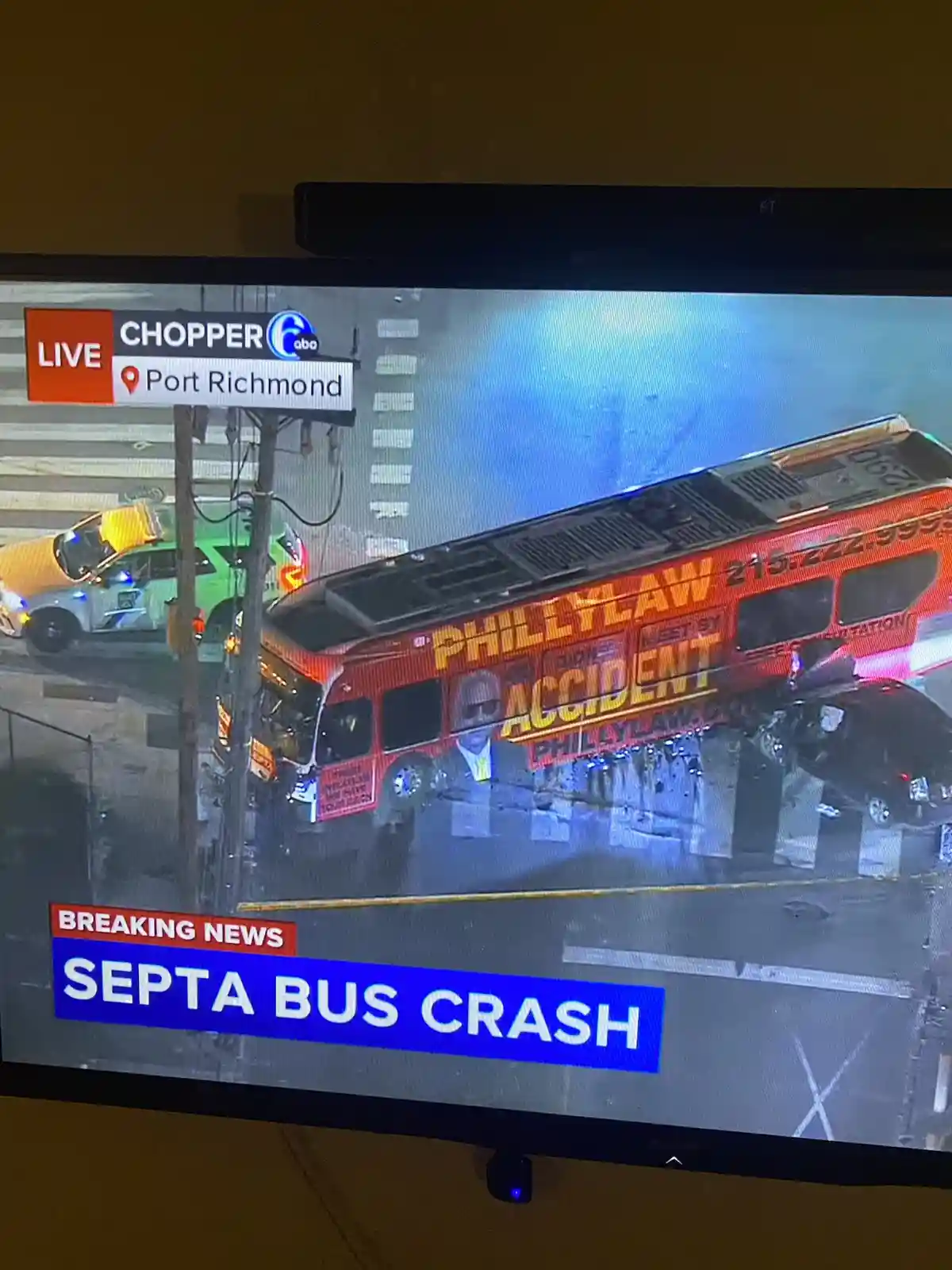 A news broadcast shows a SEPTA bus crash in Port Richmond, with a red bus tilted and damaged. The bus has an advertisement…