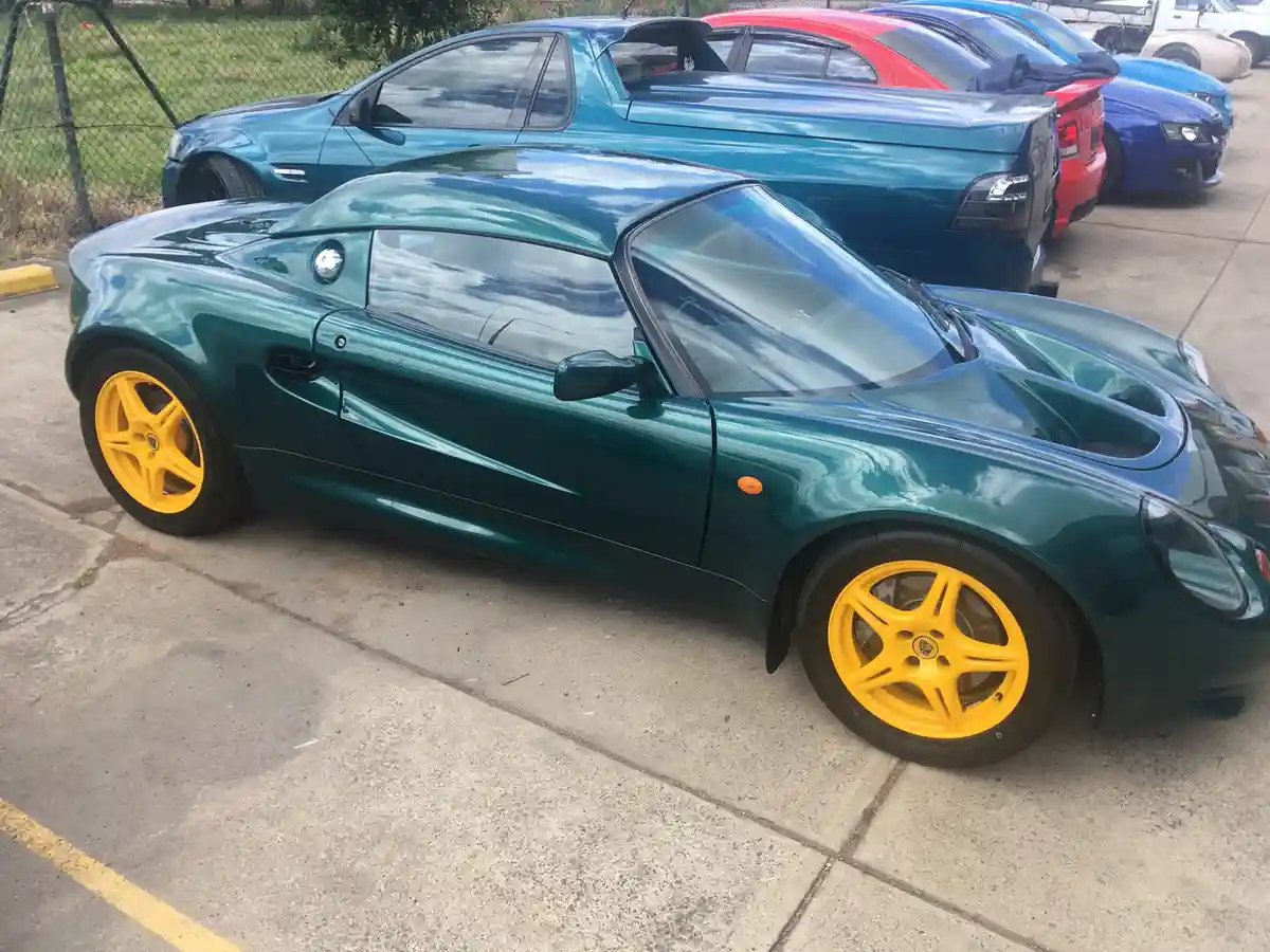 A green Lotus Elise sports car with bright yellow wheels is parked in a lot, with other cars behind it. The overlaid text…