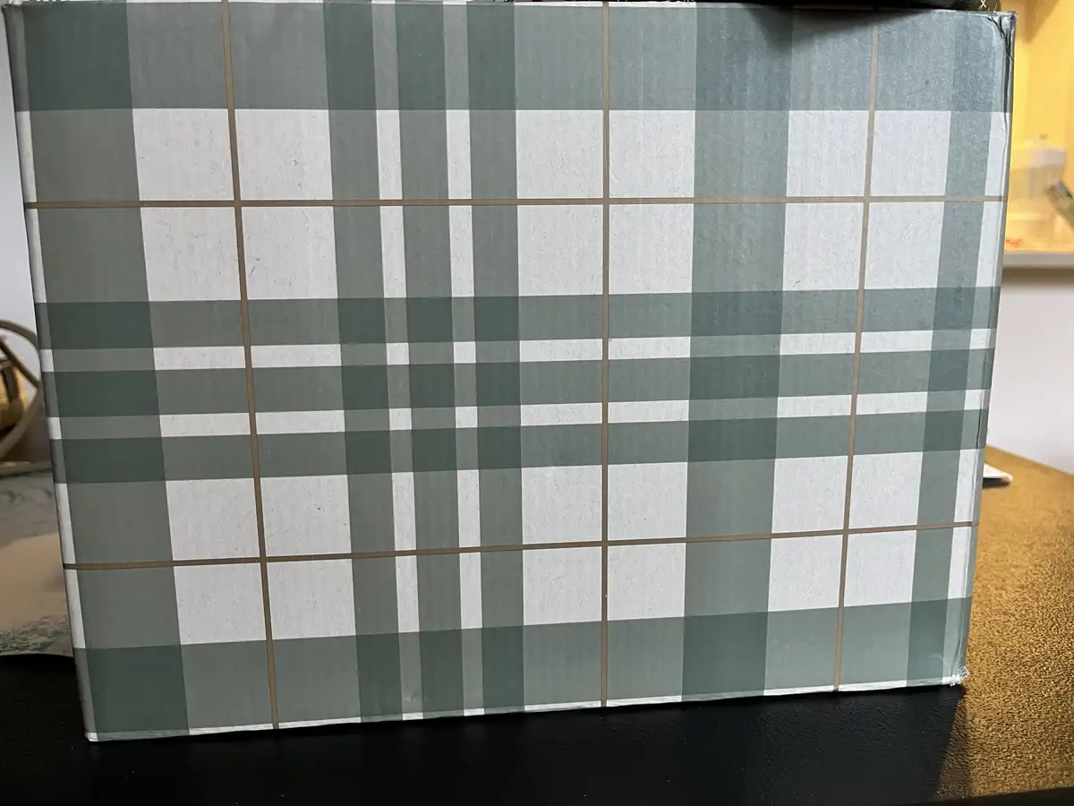 A close-up photo shows a rectangular box wrapped in green and white plaid wrapping paper with thin brown lines. The box sits…