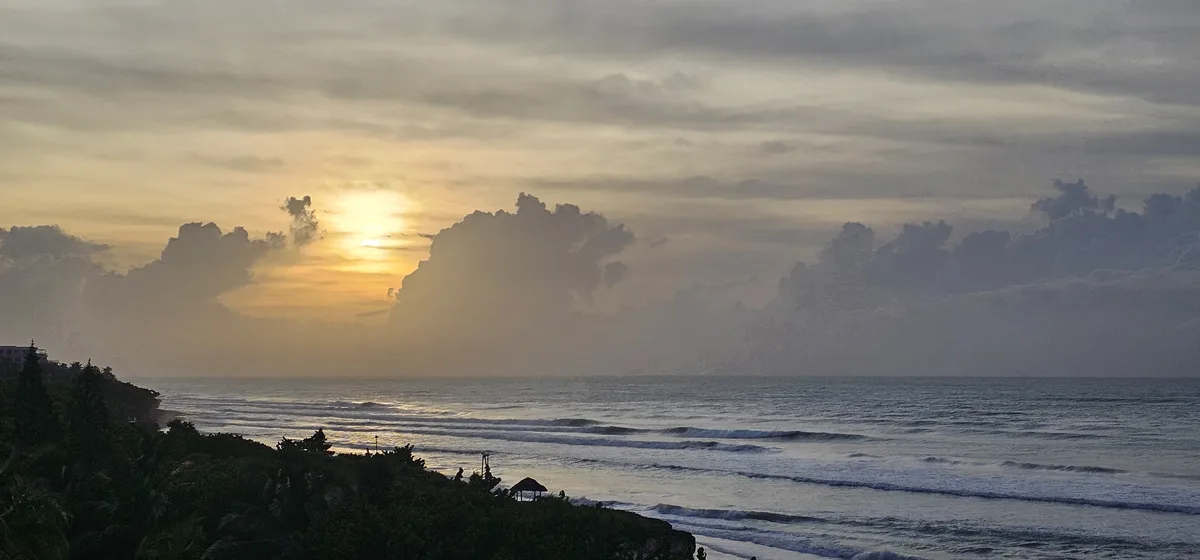 A serene sunset over the ocean with waves rolling in, silhouetted palm trees and a thatched-roof structure on the shore…