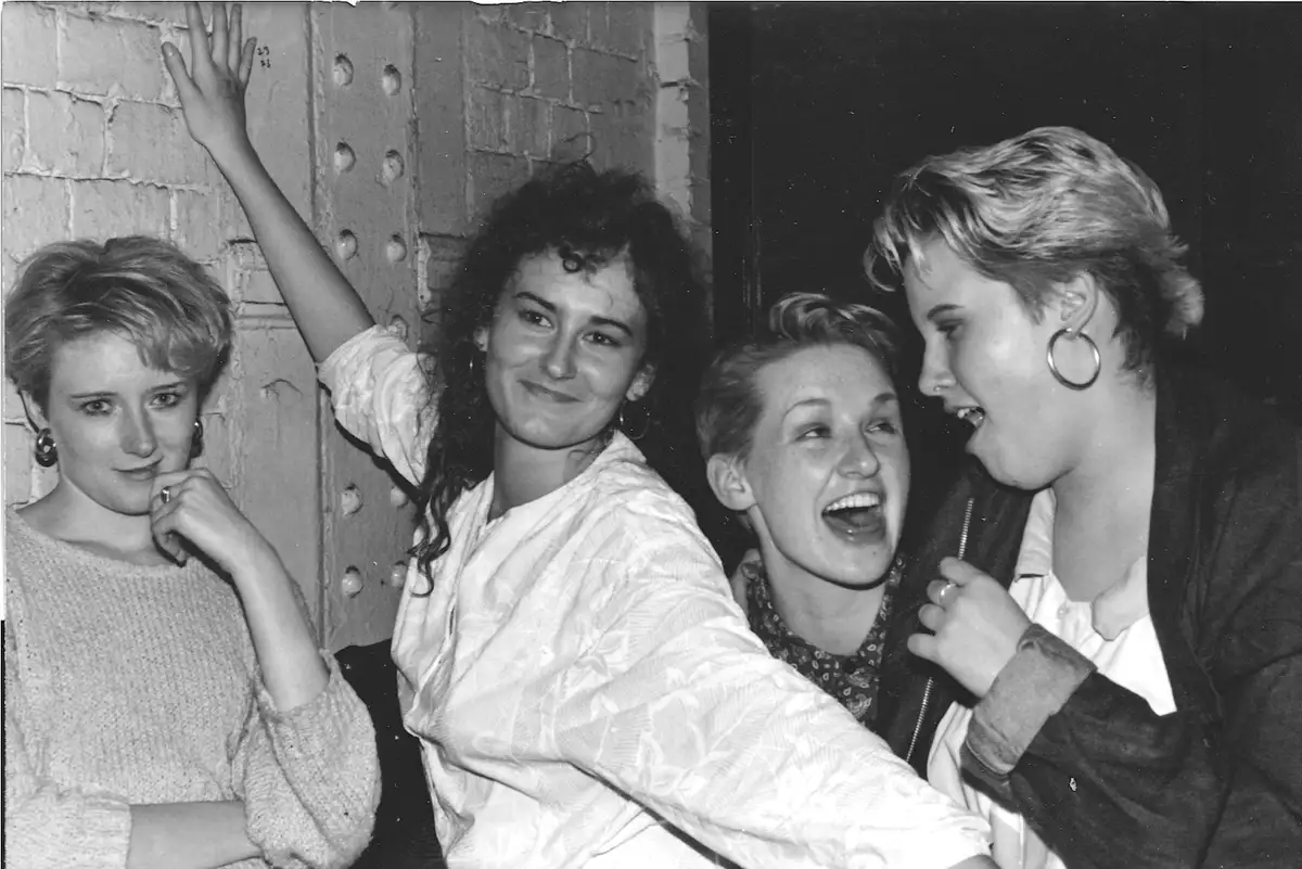 A black-and-white photo of four young women from the 1980s posing together against a brick wall. One woman on the far left…