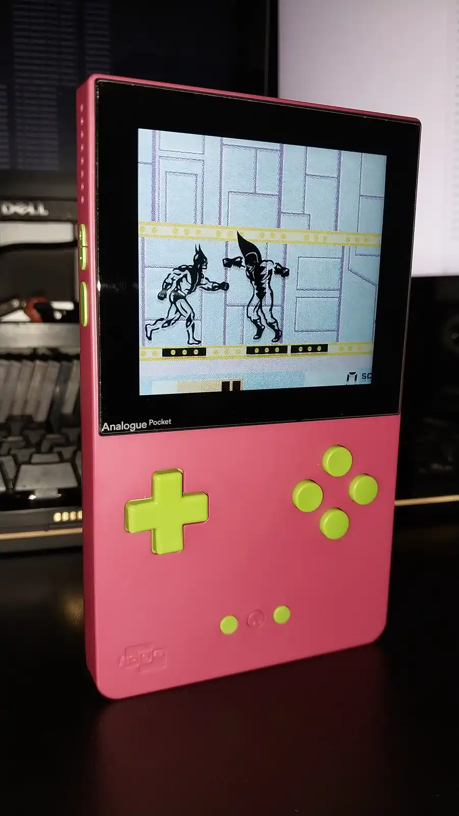 A pink Analogue Pocket handheld console displays a pixelated fighting game with two Batman-like characters in combat on a…