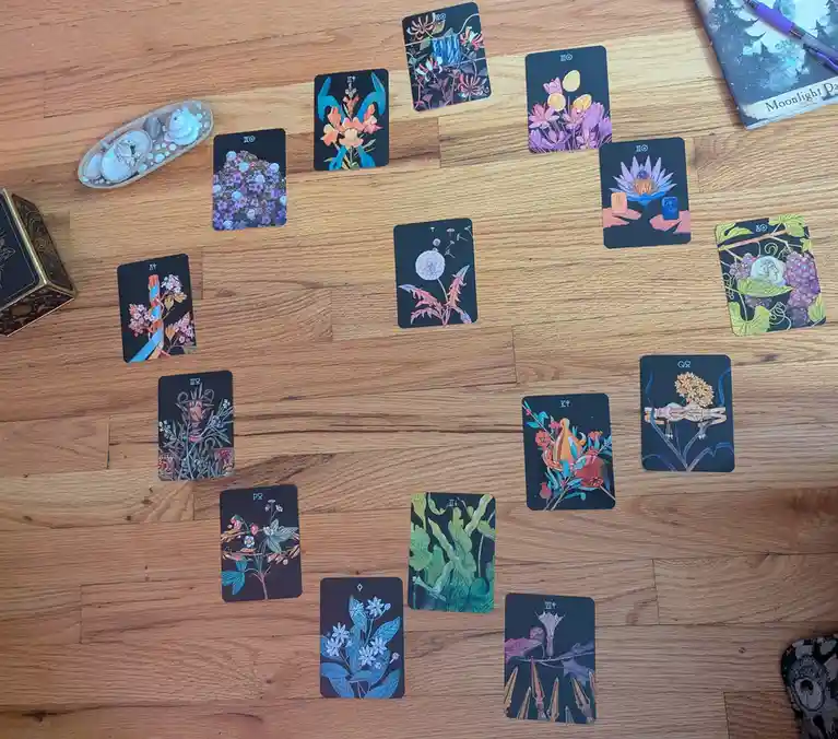 An overhead shot shows a spread of ornate, dark-backed tarot cards with colorful botanical illustrations laid out on a…