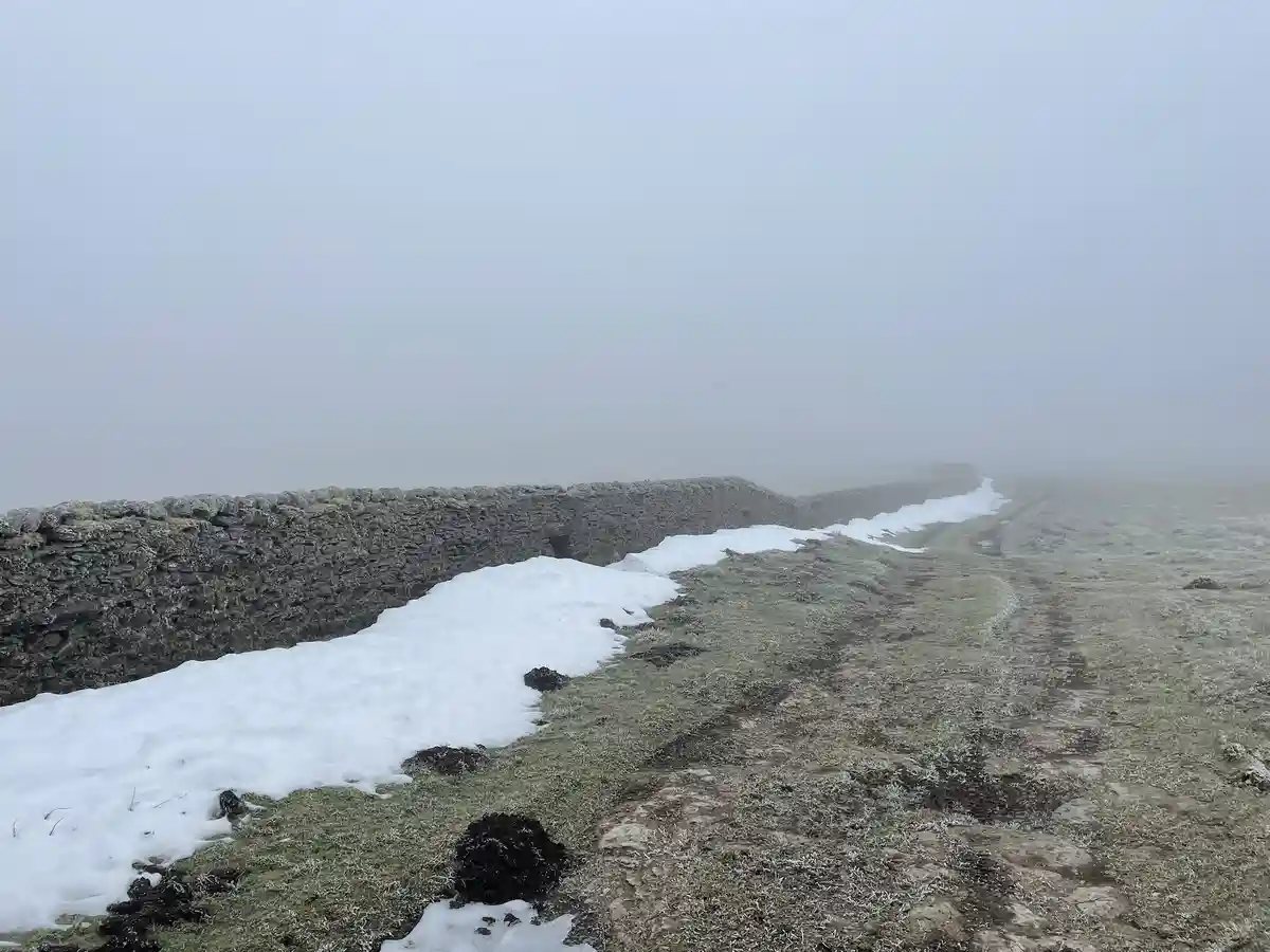 A foggy, snowy landscape shows a stone wall running alongside a muddy path, with patches of snow melting along the wall's…