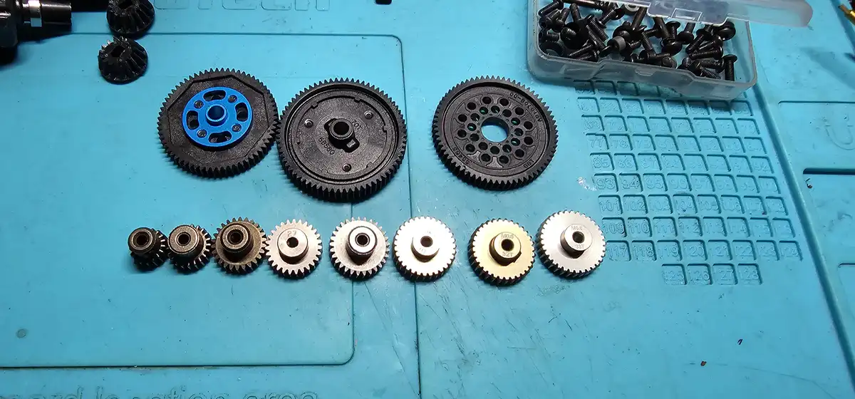 A collection of various-sized mechanical gears and cogs arranged on a blue work mat, with a container of screws nearby. The…