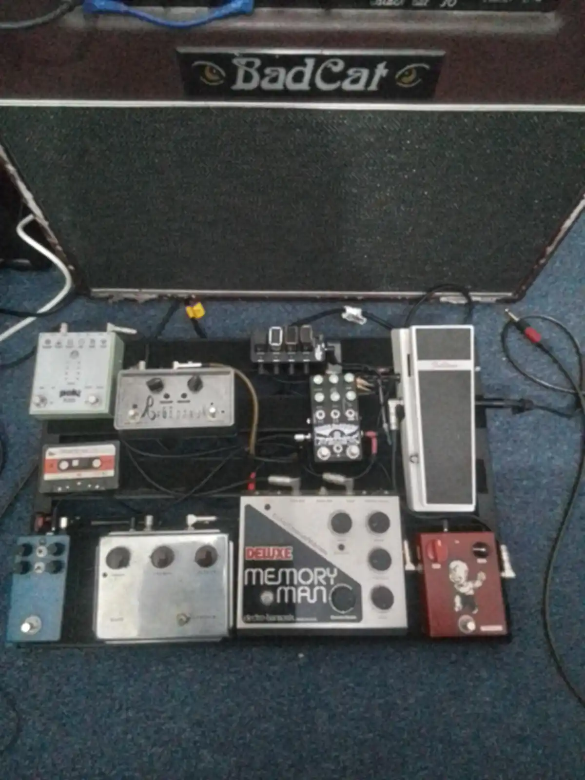 A top-down view of a densely packed guitar effects pedalboard with numerous pedals, cables, and a 'Bad Cat' amplifier head…