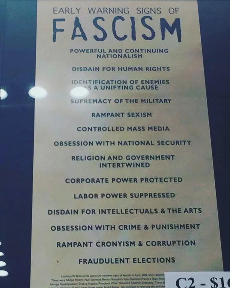 A printed poster titled 'EARLY WARNING SIGNS OF FASCISM' lists characteristics like 'POWERFUL AND CONTINUING NATIONALISM'…