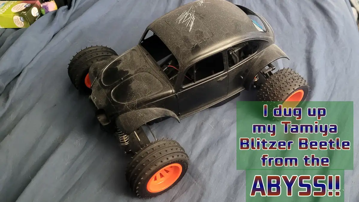 A dusty, black remote-controlled Volkswagen Beetle toy with spiked tires and orange hubs rests on a blue fabric surface…