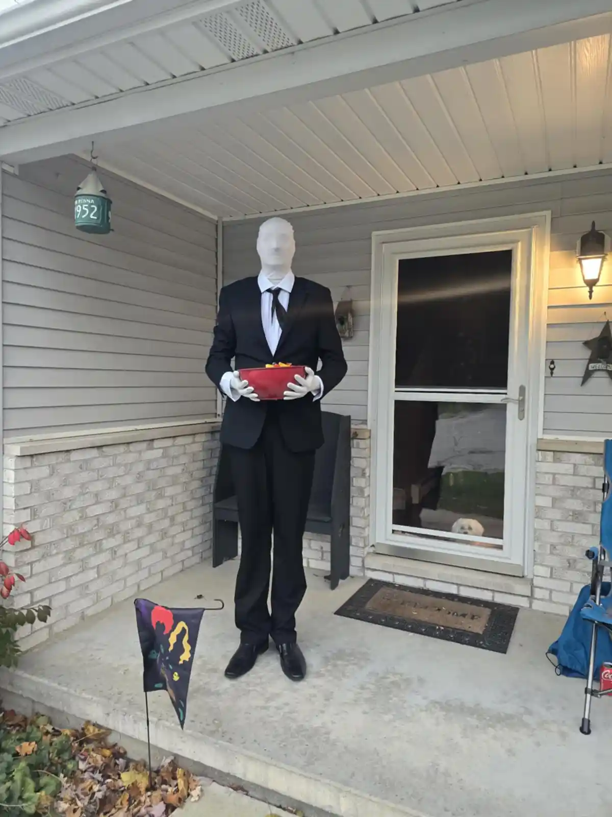 A mannequin with a blank white head, dressed in a black suit and tie, stands on a front porch holding a red bowl of food…