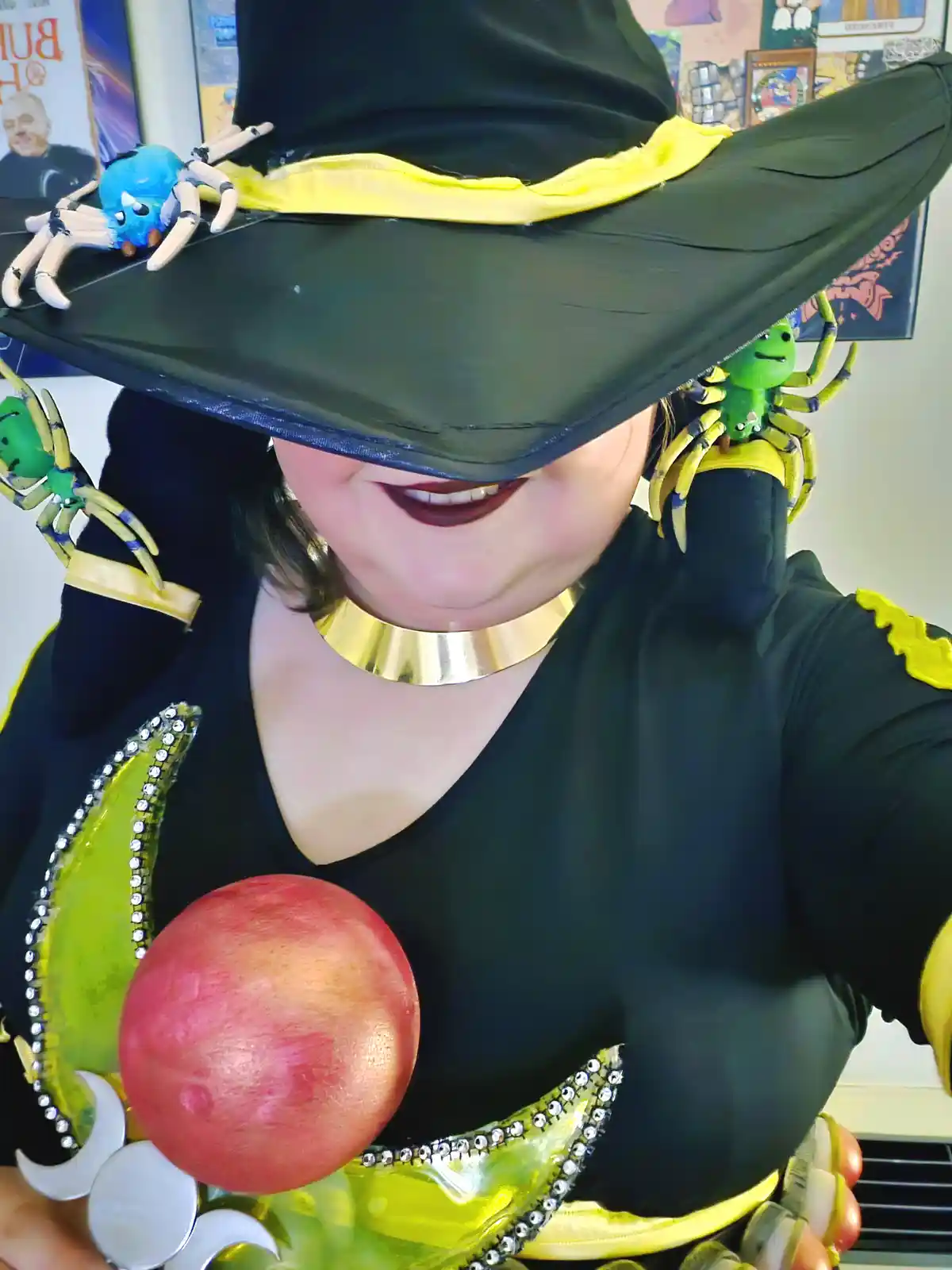 A person in a witch costume with a large black hat and gold collar is smiling while holding a red orb. Several colorful…