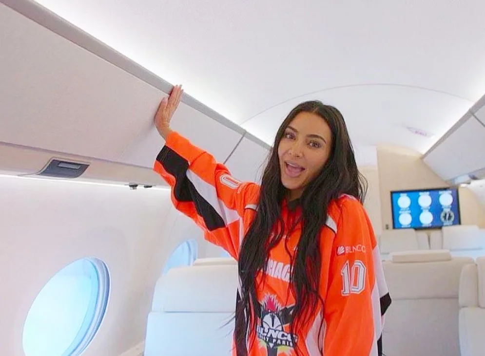 Kim Kardashian is inside a private jet, wearing an orange and black sports jersey with the number 10 and 'BLNCO' logo…