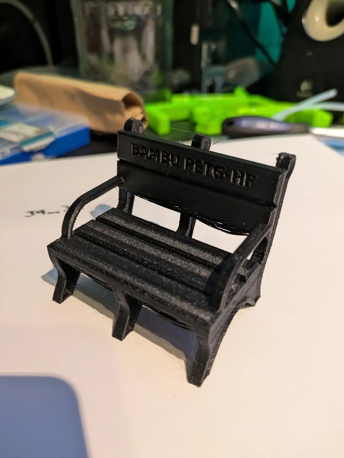 A small, black 3D-printed miniature bench sits on a white surface, with visible layer lines and a slightly rough texture…