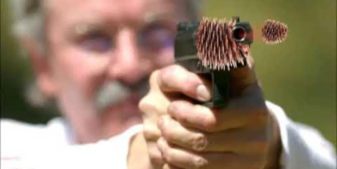 A blurry image of an older man with a mustache holding a handgun pointed forward. The gun's barrel and the bullet are…