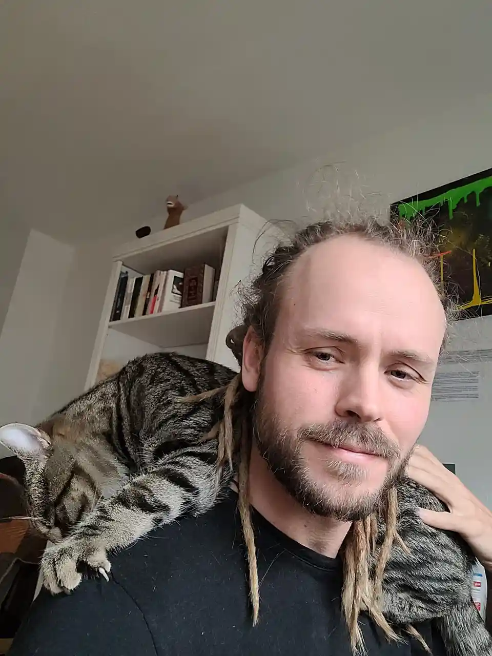 A man with dreadlocks and a beard takes a selfie, looking directly at the camera with a slight smirk. A tabby cat is curled…