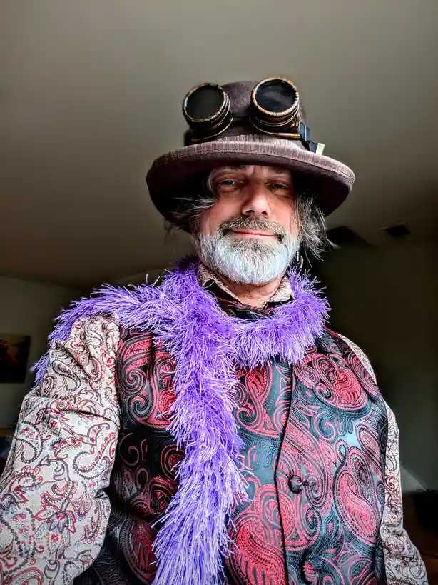 A man with a white beard and mustache wears a brown top hat with goggles on top, a purple feather boa, and a colorful…