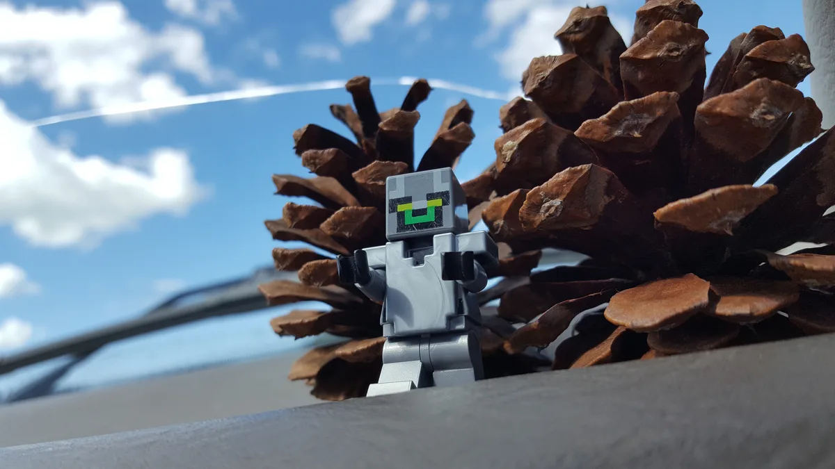 A small gray Minecraft creeper LEGO minifigure stands in front of a large brown pinecone, with a blue sky and clouds visible…