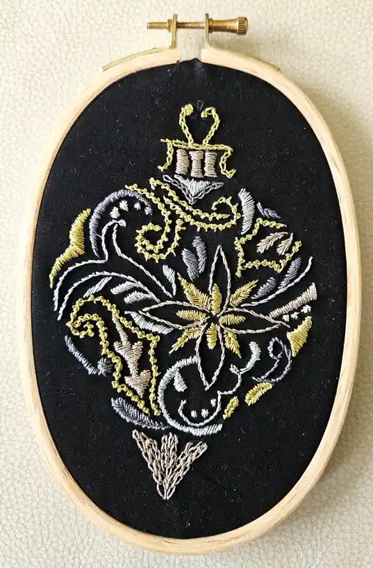 An embroidery hoop holds a black fabric with intricate, ornate stitching in gold, silver, and beige, forming a symmetrical…