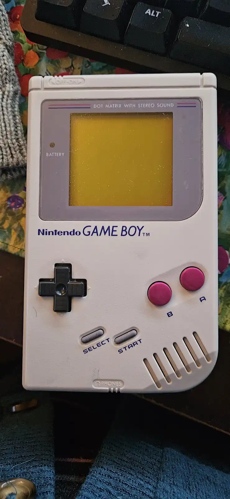 A close-up photo of a white Nintendo Game Boy with a yellow-tinted screen, pink A and B buttons, and a black directional…