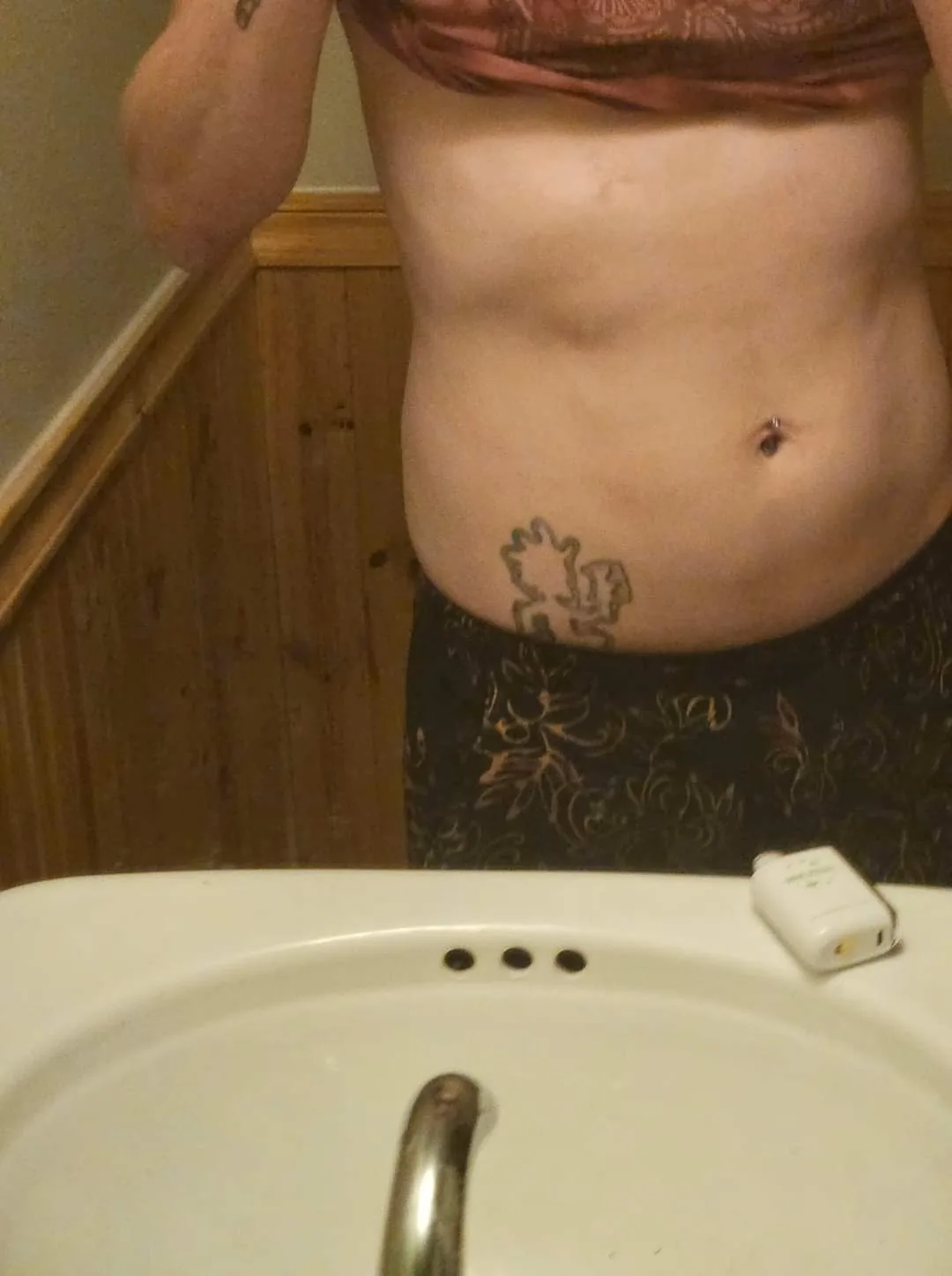 A person stands in front of a bathroom mirror, shirtless, showing their midsection with a visible tattoo near the hip and a…
