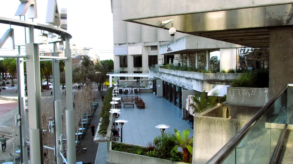 An outdoor urban plaza with concrete architecture, tables, benches, and sparse people, featuring a visible security camera…