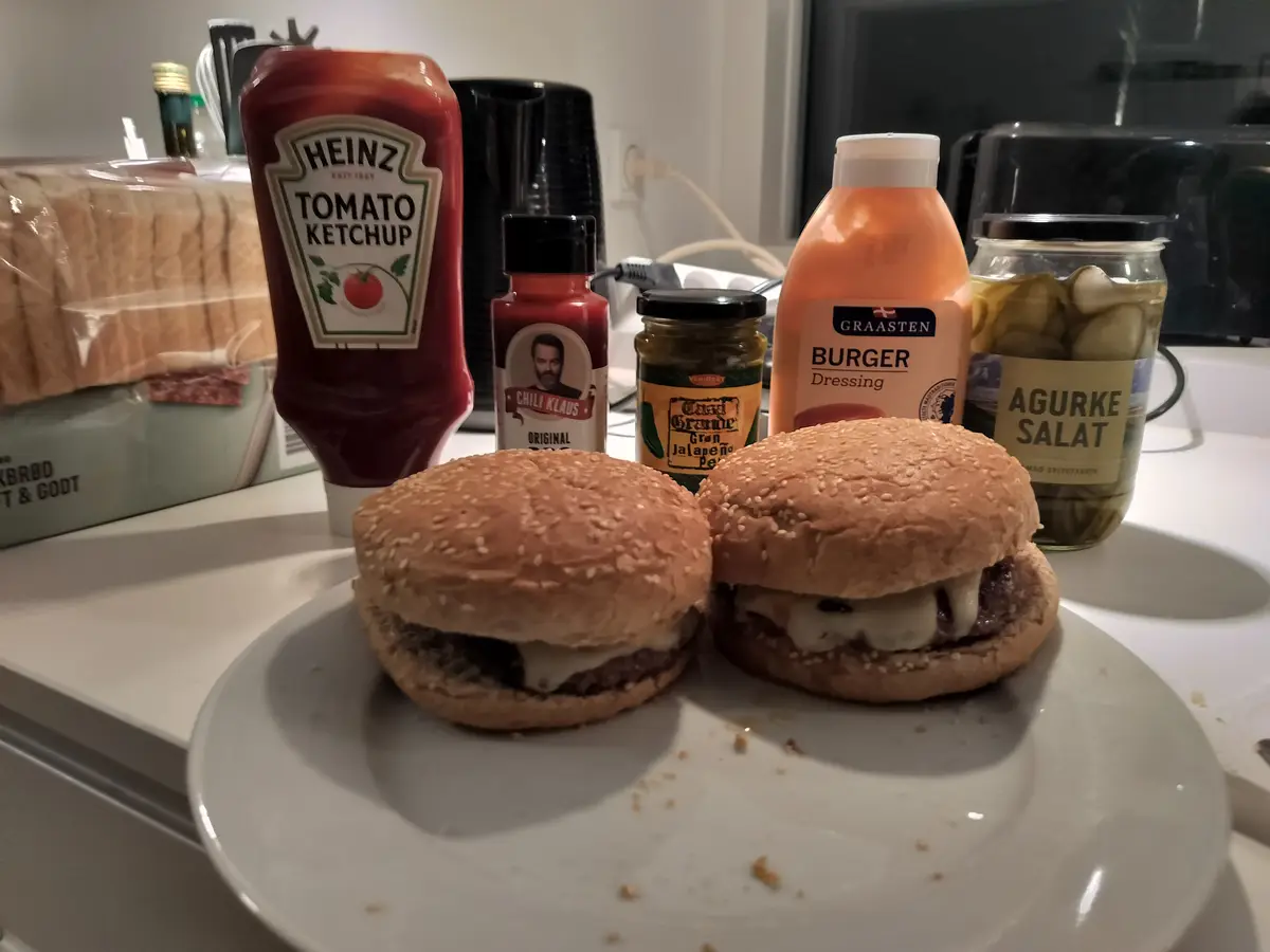 Two plain cheeseburgers sit on a white plate, surrounded by various condiment bottles and jars including Heinz ketchup…
