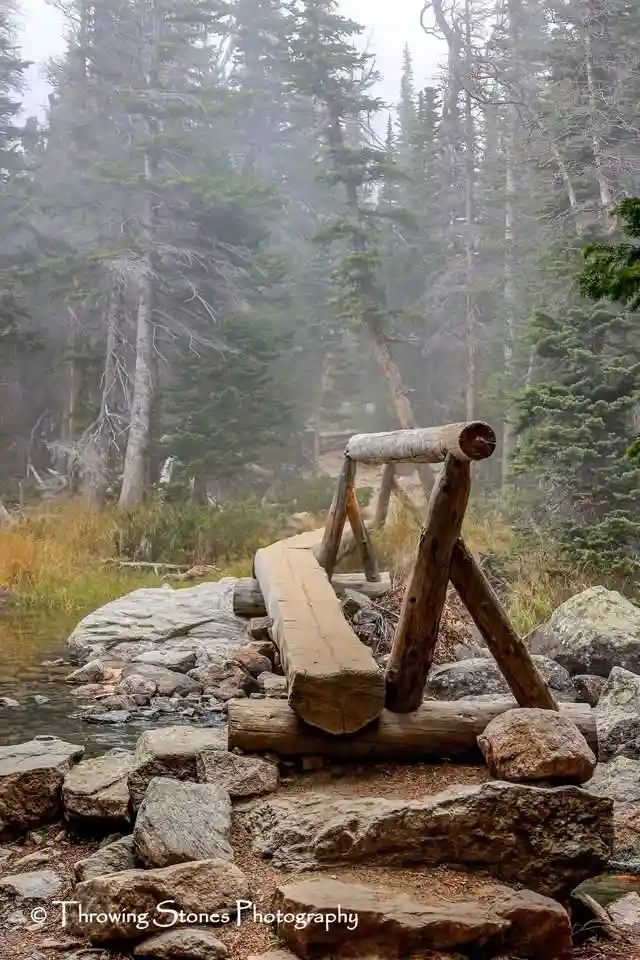A rustic wooden bridge made of logs and rocks spans a small stream in a foggy forest with tall pine trees. The overlaid text…
