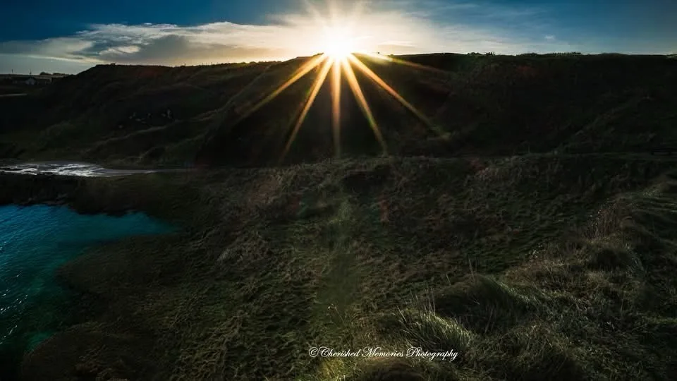 A scenic photo of a sun setting or rising over a grassy cliffside with ocean water visible below, featuring prominent sun…