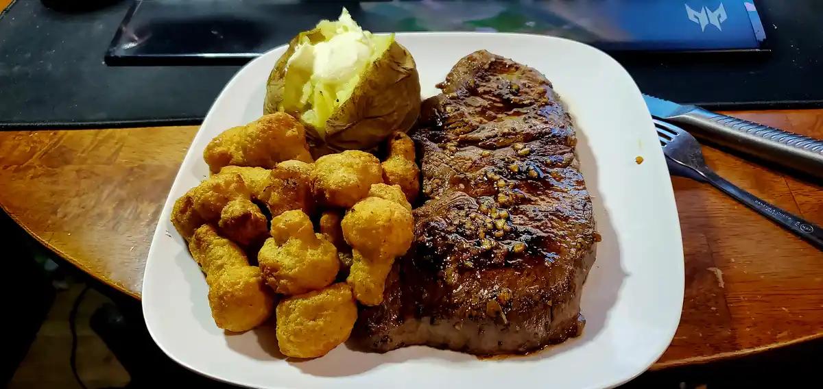 A white plate holds a large, seared steak with a glossy sauce, golden-brown tater tots, and a baked potato topped with…