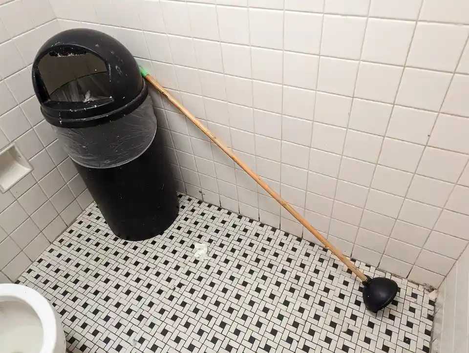A black trash can with a plastic liner sits in a tiled bathroom corner next to a plunger with a wooden handle and black…