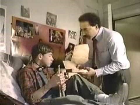 a man in a plaid shirt sits in bed holding a glass bottle while another man in a collared shirt and tie stands beside him…