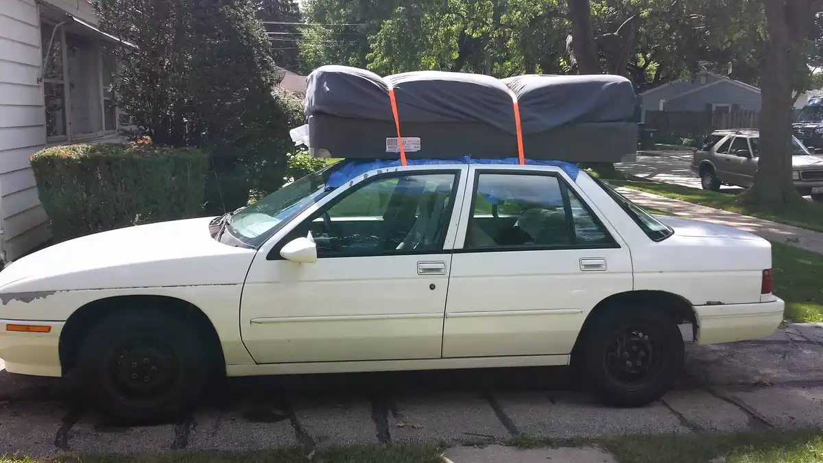 A white sedan is parked on a residential street with three mattresses strapped to its roof using orange straps and blue…