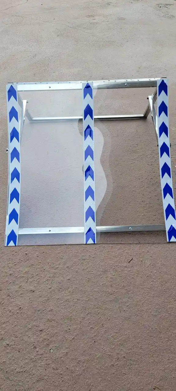 A transparent plastic barrier with blue and white chevron warning tape on its vertical edges lies on a paved surface. The…