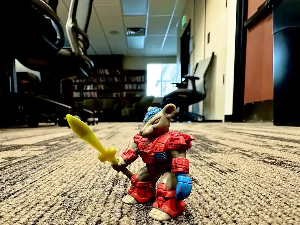 A small, red-armored mouse toy holding a yellow sword stands on a carpeted office floor, with office chairs and bookshelves…