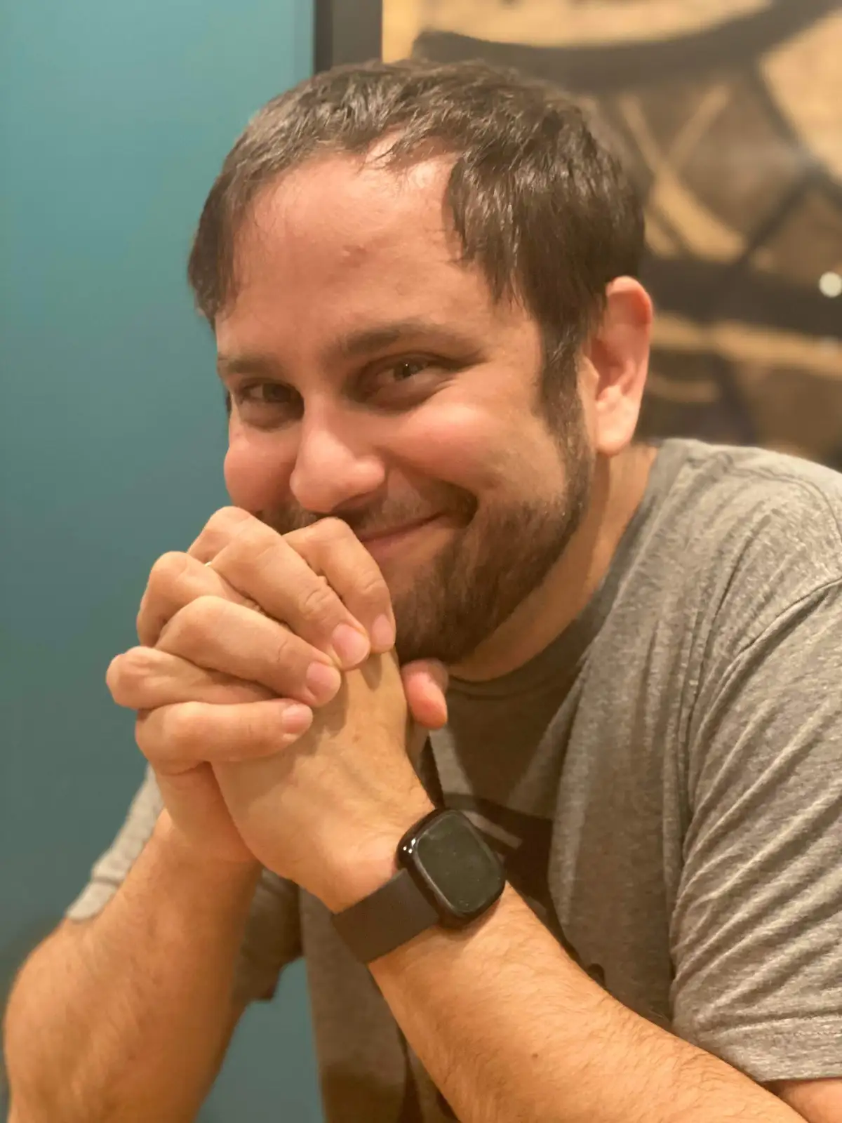 A man with dark hair and a beard, wearing a gray t-shirt and a black smartwatch, smiles while resting his clasped hands…
