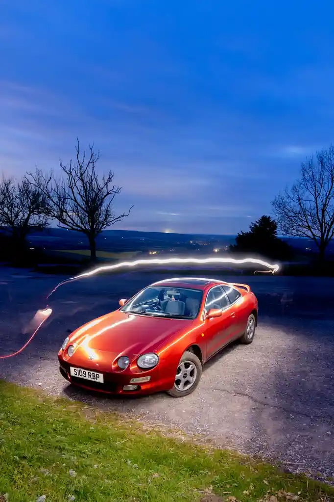 A red Toyota Celica sits parked on a gravel road at dusk, with a long-exposure light trail resembling a glowing, serpentine…