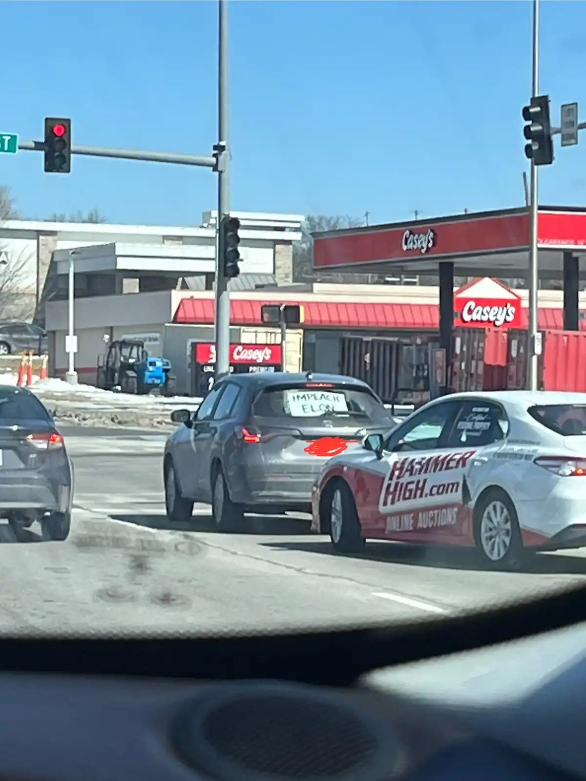 A gray SUV is stopped at a red traffic light with a handwritten sign in its rear window that reads 'IMPEACH ELON'. The SUV…