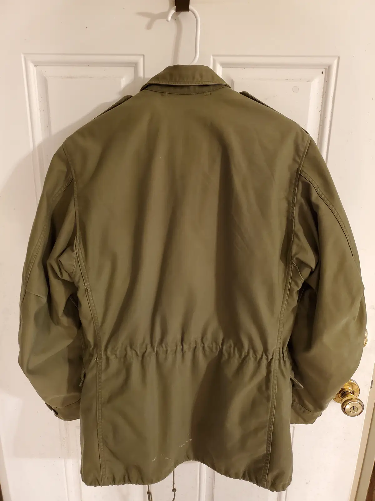 An olive green military-style jacket hangs on a white plastic hanger against a white interior door, viewed from the back…