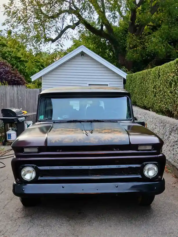 A front-facing photo of a vintage maroon Chevrolet truck parked in a driveway, with a white shed and green hedge in the…