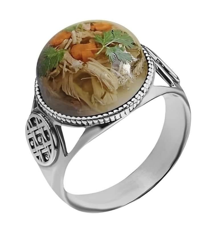 A silver ring with a large, round, clear stone set in the center, which contains a preserved, edible-looking soup with…