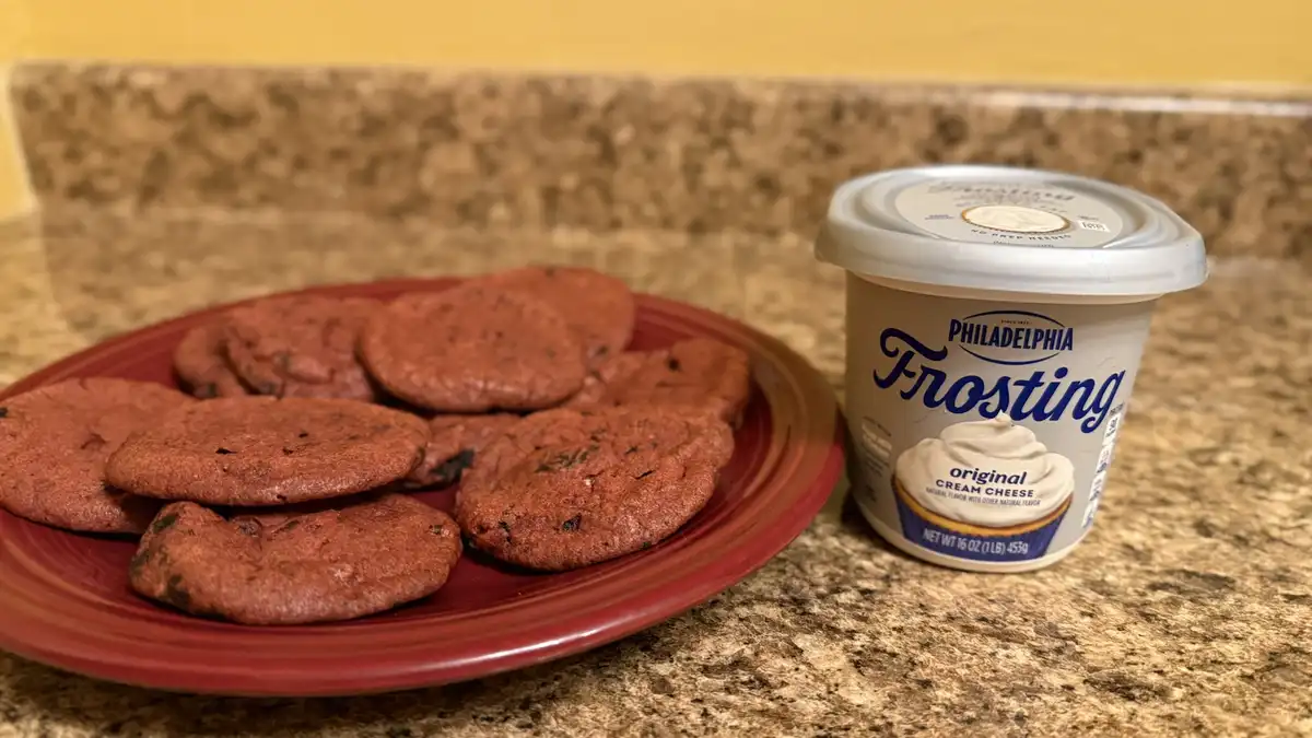 A red plate of red velvet cookies sits next to a tub of Philadelphia Original Cream Cheese Frosting on a granite countertop…
