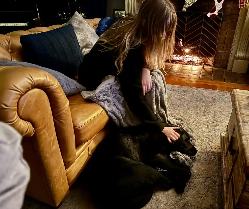 A person with long blonde hair sits on a tan leather couch, petting a black dog lying on a rug in front of them. A fireplace…