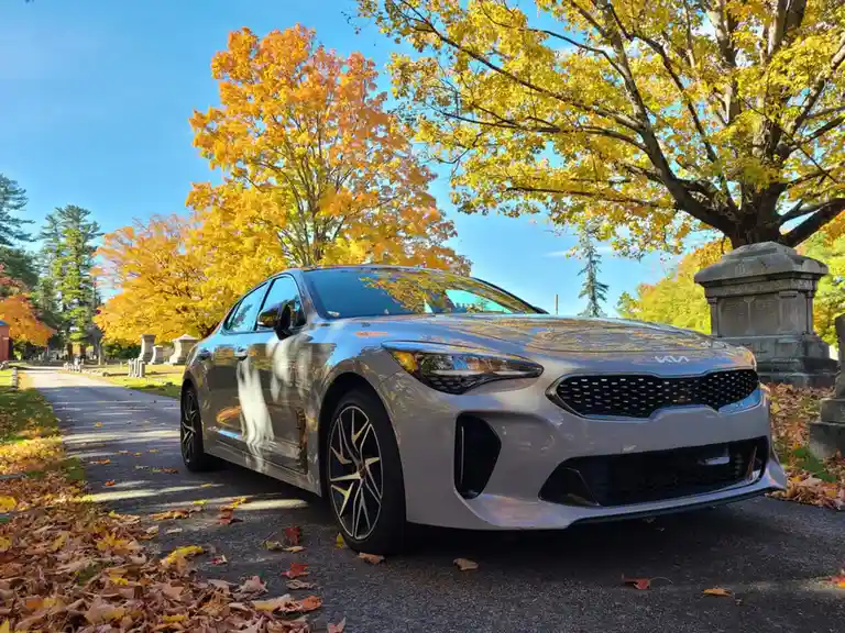 A silver Kia Stinger sedan is parked on a paved path in a cemetery during autumn, surrounded by vibrant yellow trees and…