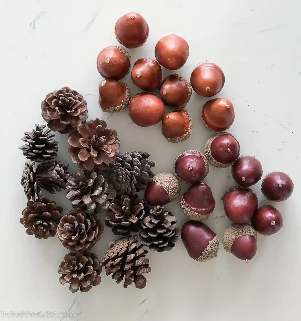 An overhead shot displays a collection of pinecones and acorns arranged on a white marble surface. The pinecones are…
