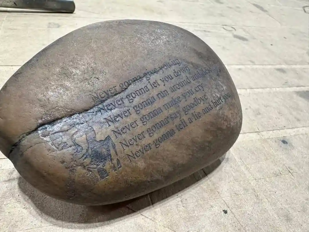 A smooth, rounded stone with the lyrics of 'Never Gonna Give You Up' engraved on its surface, resting on a wooden workbench…