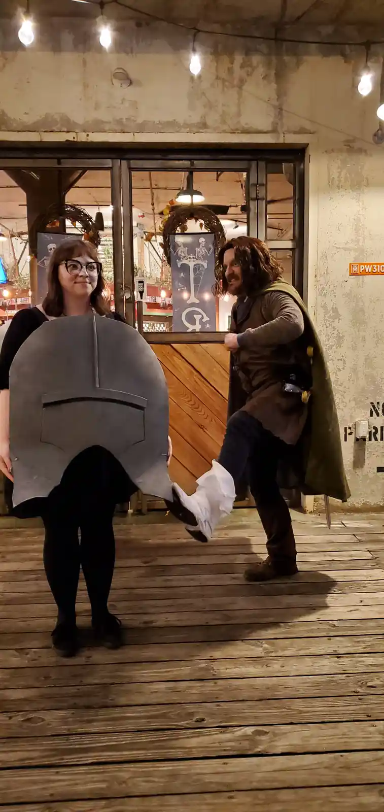 A woman wearing a large, grey, helmet-shaped shield costume stands still while a man in a fantasy costume kicks his leg up…