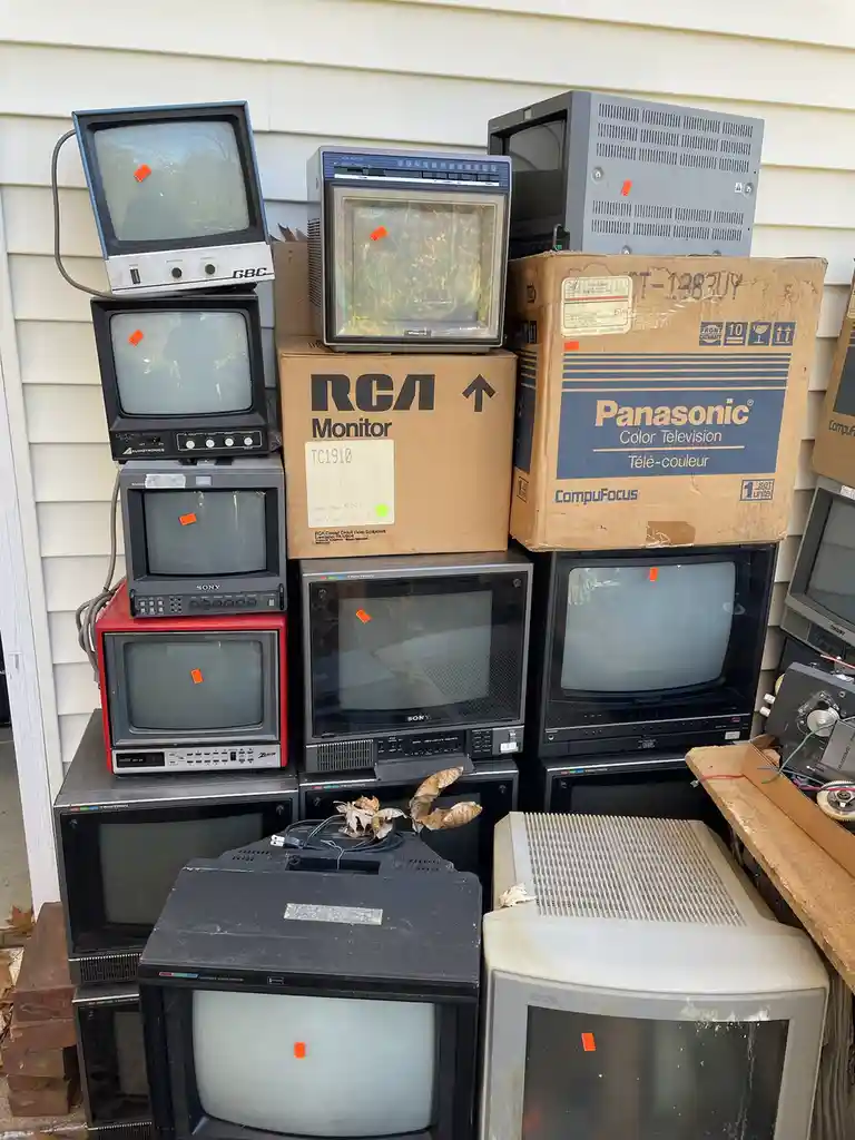 A large pile of old, mostly off-brand and vintage CRT televisions and monitors stacked outdoors against a house wall, many…