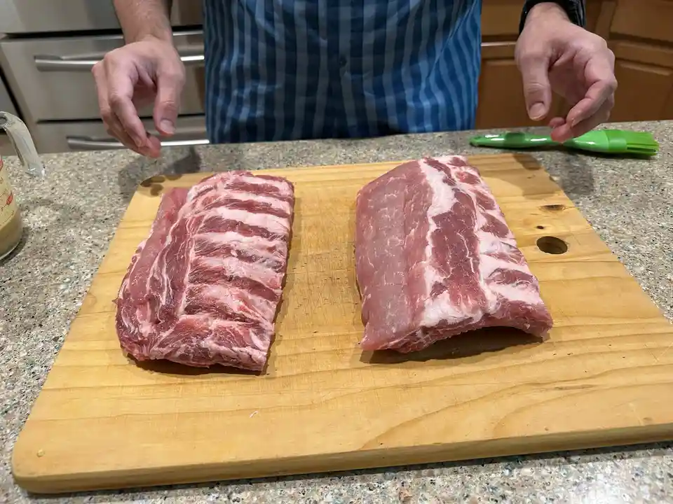 A person in a blue and white striped shirt stands behind a wooden cutting board on a kitchen counter, with two raw pork rib…