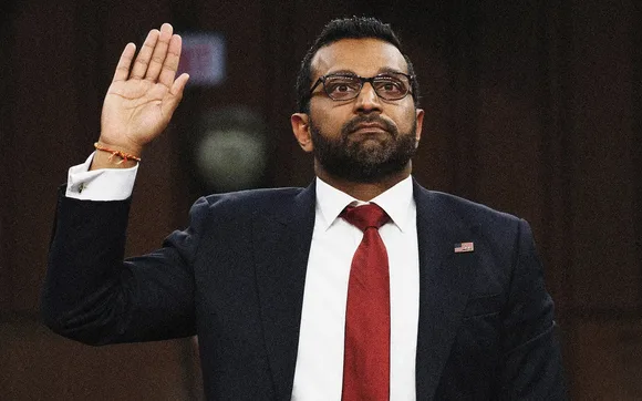 A man in a dark suit, white shirt, and red tie raises his right hand in an oath-taking gesture. He wears glasses and has a…