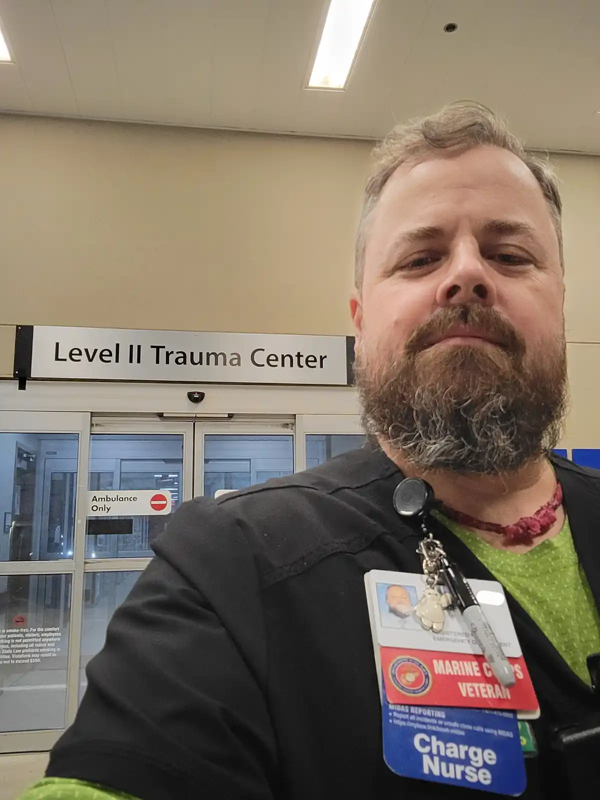 A man with a beard and a lanyard with multiple badges, including 'Charge Nurse' and 'Marine Corps Veteran', takes a selfie…